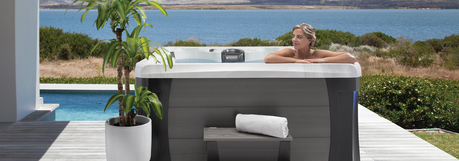 Woman relaxing in a Marquis Broadway Elite hot tub on a modern deck overlooking a pool and scenic landscape.
