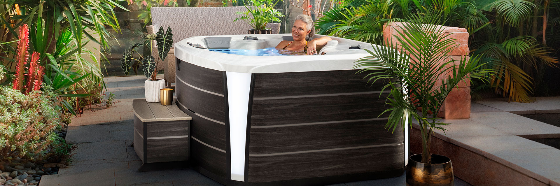 Woman relaxing in a Marquis Crown Spirit hot tub surrounded by lush garden plants in an outdoor spa setting.
