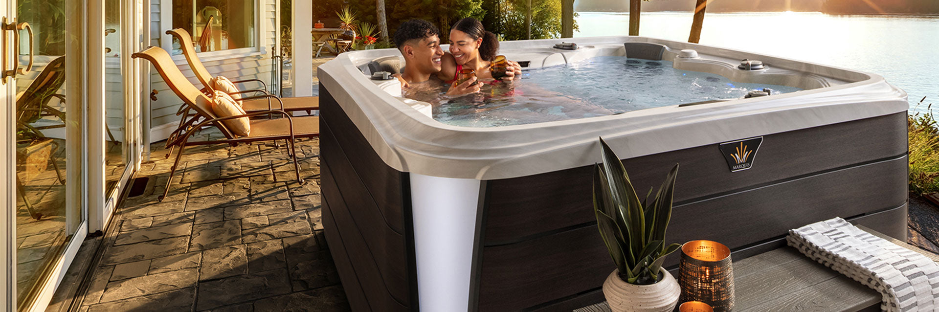 Couple relaxing in a Marquis Destiny hot tub on a lakeside patio at sunset.
