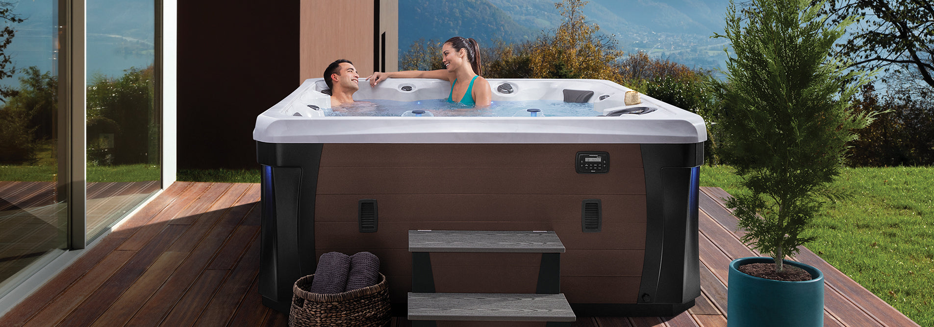 Couple relaxing in a Marquis Hollywood Elite hot tub on a wooden deck with mountain and garden views.