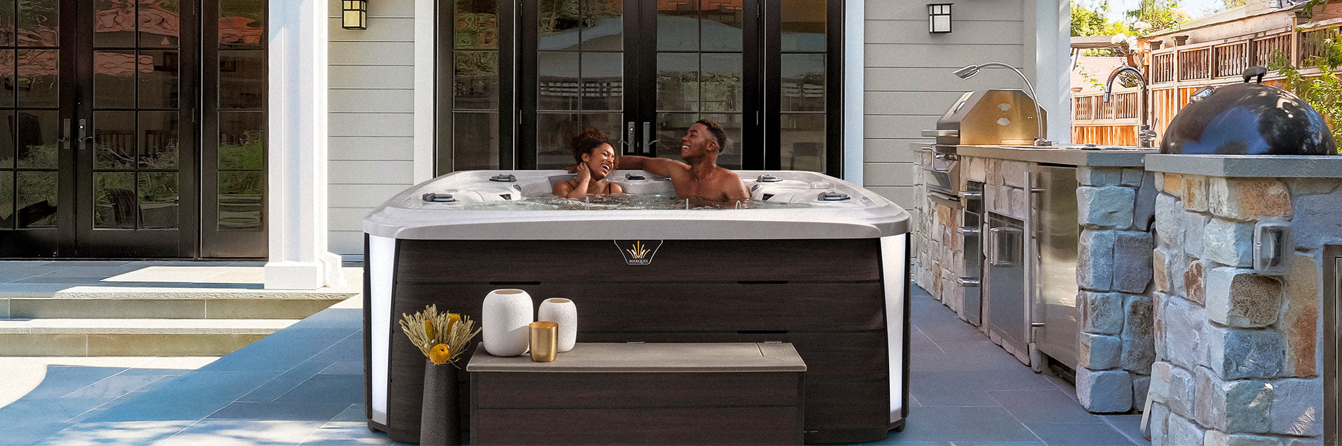 Couple relaxing in the Marquis Crown Series Euphoria hot tub installed beside an outdoor kitchen in a modern backyard setting.