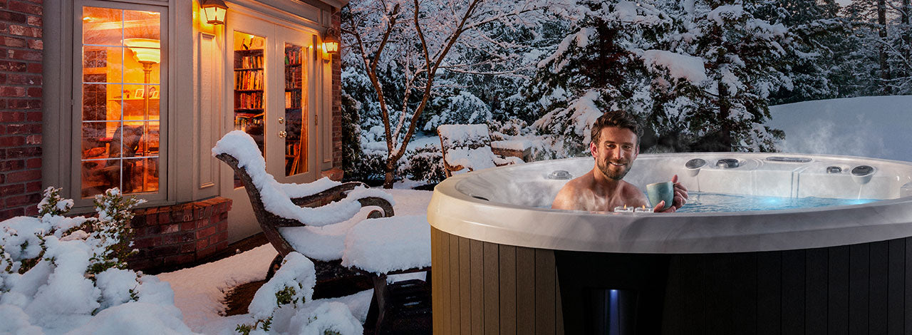 Man relaxing in a Monaco Elite hot tub outdoors in winter with snow-covered trees and a warm-lit home in the background.
