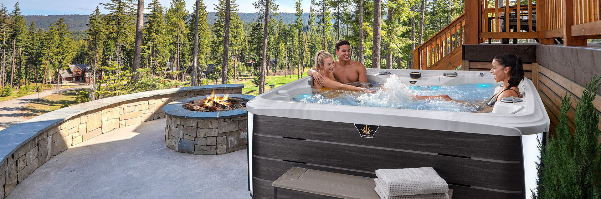 Outdoor lifestyle scene featuring people relaxing in a Marquis Summit hot tub beside a stone fire pit in a forest setting.