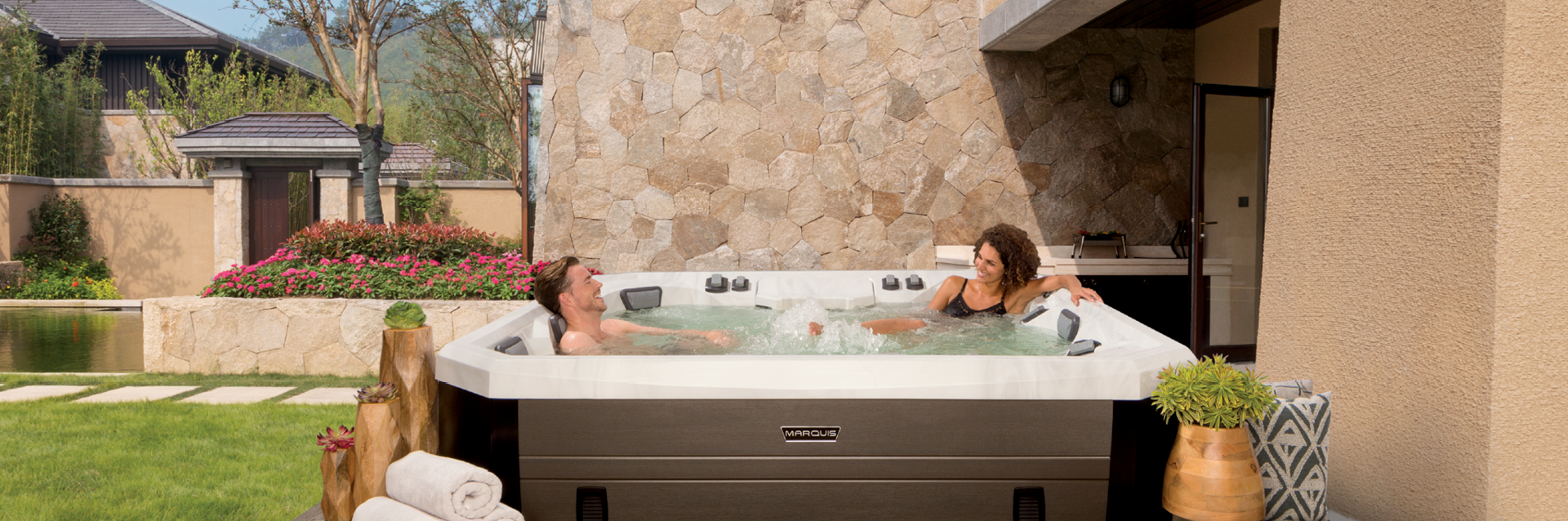 Couple relaxing in a Marquis Vector21 Series V94L hot tub in a landscaped backyard with stone walls and greenery.