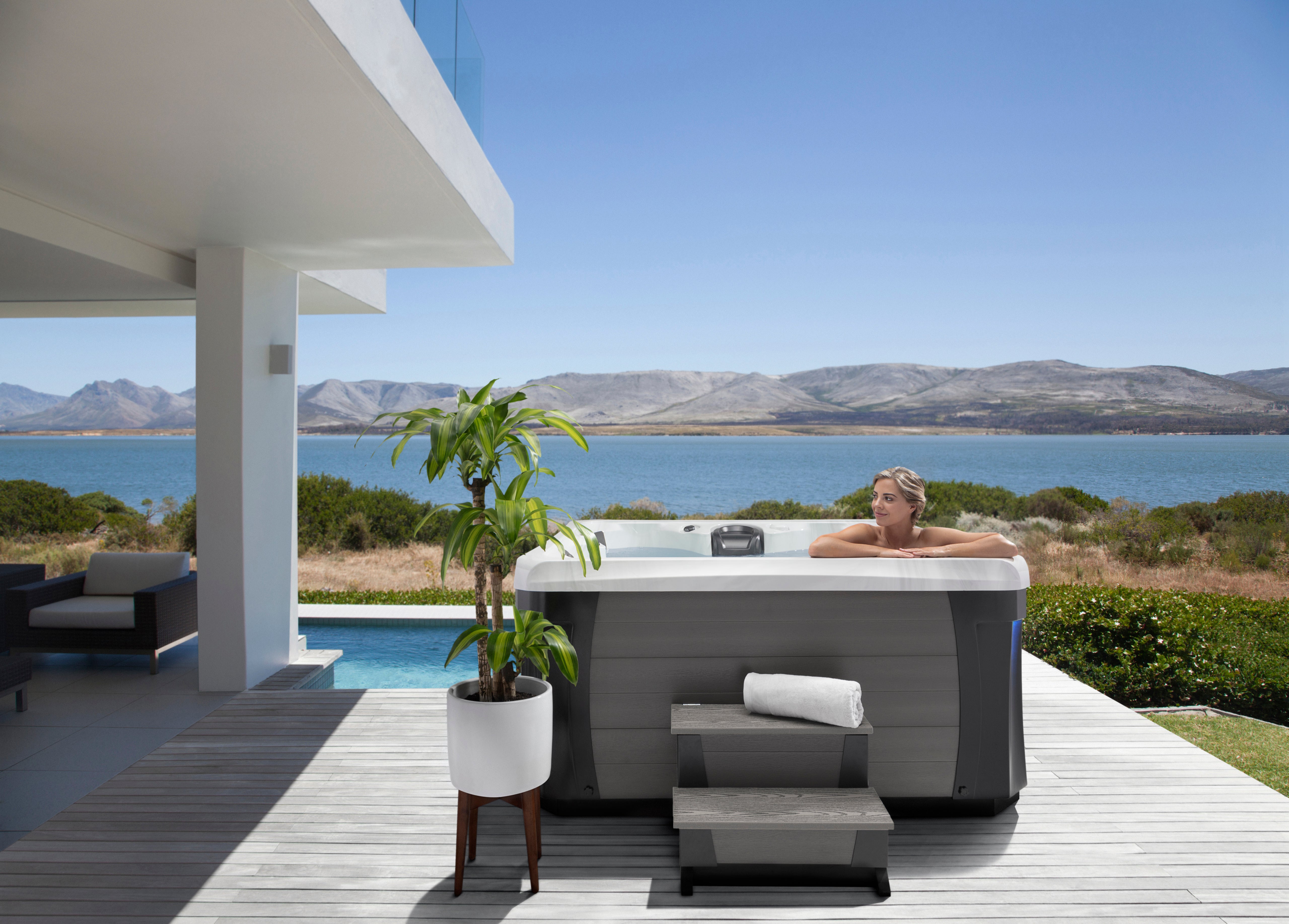Woman relaxing in an outdoor hot tub on a modern patio overlooking a lake and mountains.