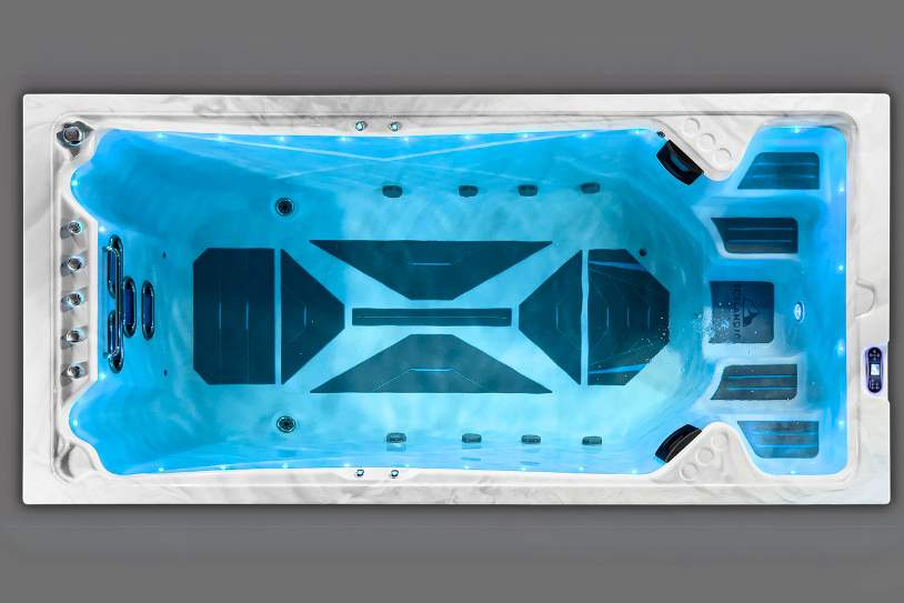Top-down view of a large illuminated swim spa filled with still blue water.