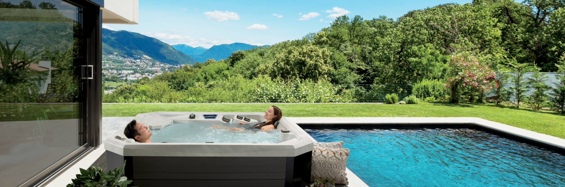 Couple relaxing in a Vector21 Series V84L hot tub on a patio beside a swimming pool with mountains and greenery.