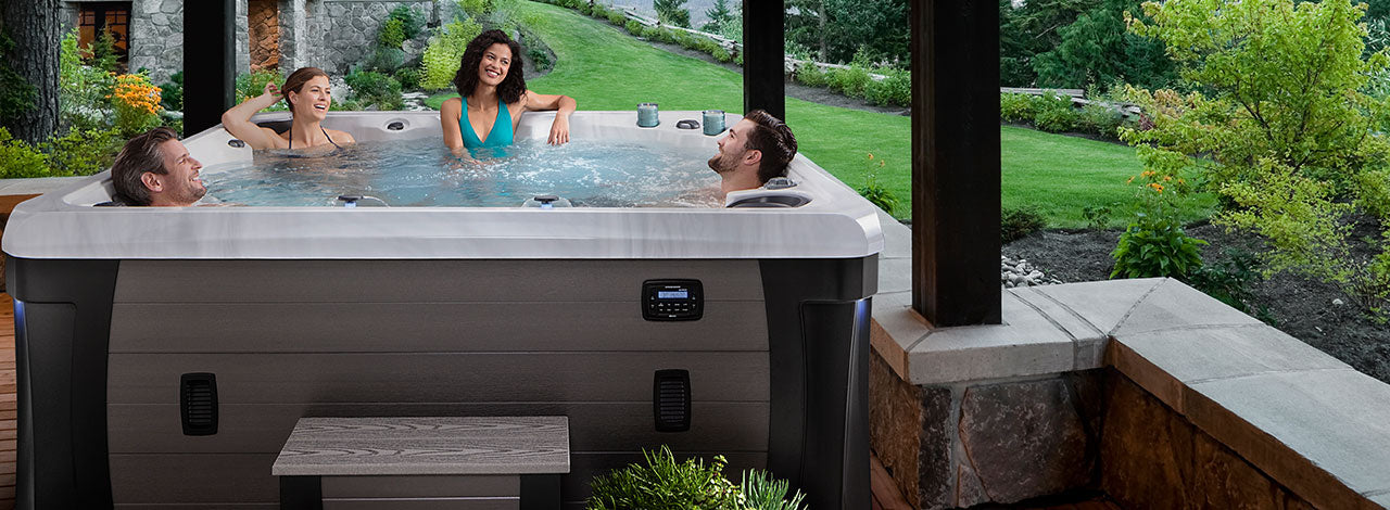 Group of people relaxing in a Marquis Woodstock Elite hot tub on a covered patio overlooking a landscaped backyard.